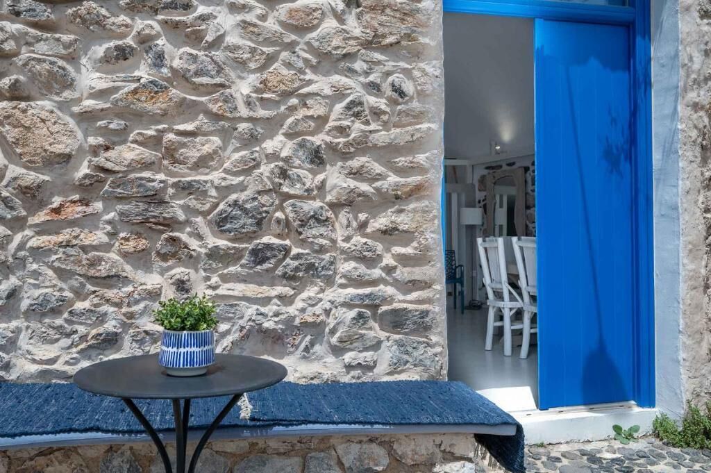 Un muro de piedra con una puerta azul, una mesa redonda con una planta en maceta y un vistazo a muebles blancos en el interior.