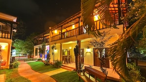 A well-lit hotel with a covered outdoor seating area, a pathway leading to the entrance, and lush greenery. - Amour Resort (Vagator)