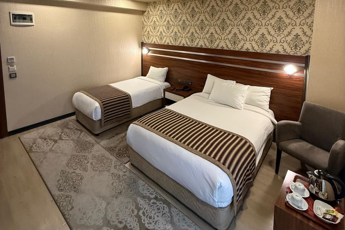 A hotel room with a large bed, two smaller beds, a wooden headboard, a patterned rug, a chair, and a small table with a tray.