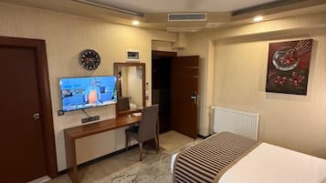 A hotel room with a bed, a desk with a chair, a television, a clock, and a radiator.