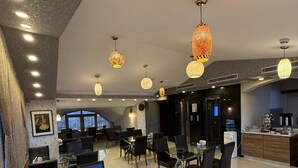 Un hall d’hôtel moderne avec des tables et des chaises en verre, des luminaires décoratifs suspendus et un espace cuisine visible.