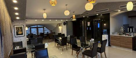 Un moderno vestíbulo de hotel con mesas y sillas de vidrio, lámparas decorativas colgantes y una zona de cocina a la vista.