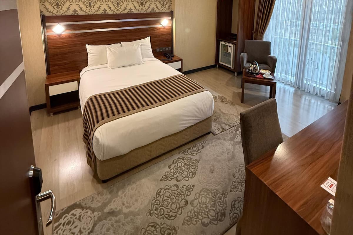 A hotel room with a large bed, a desk, a chair, a small table with a lamp, and a view of the outdoors through a window.