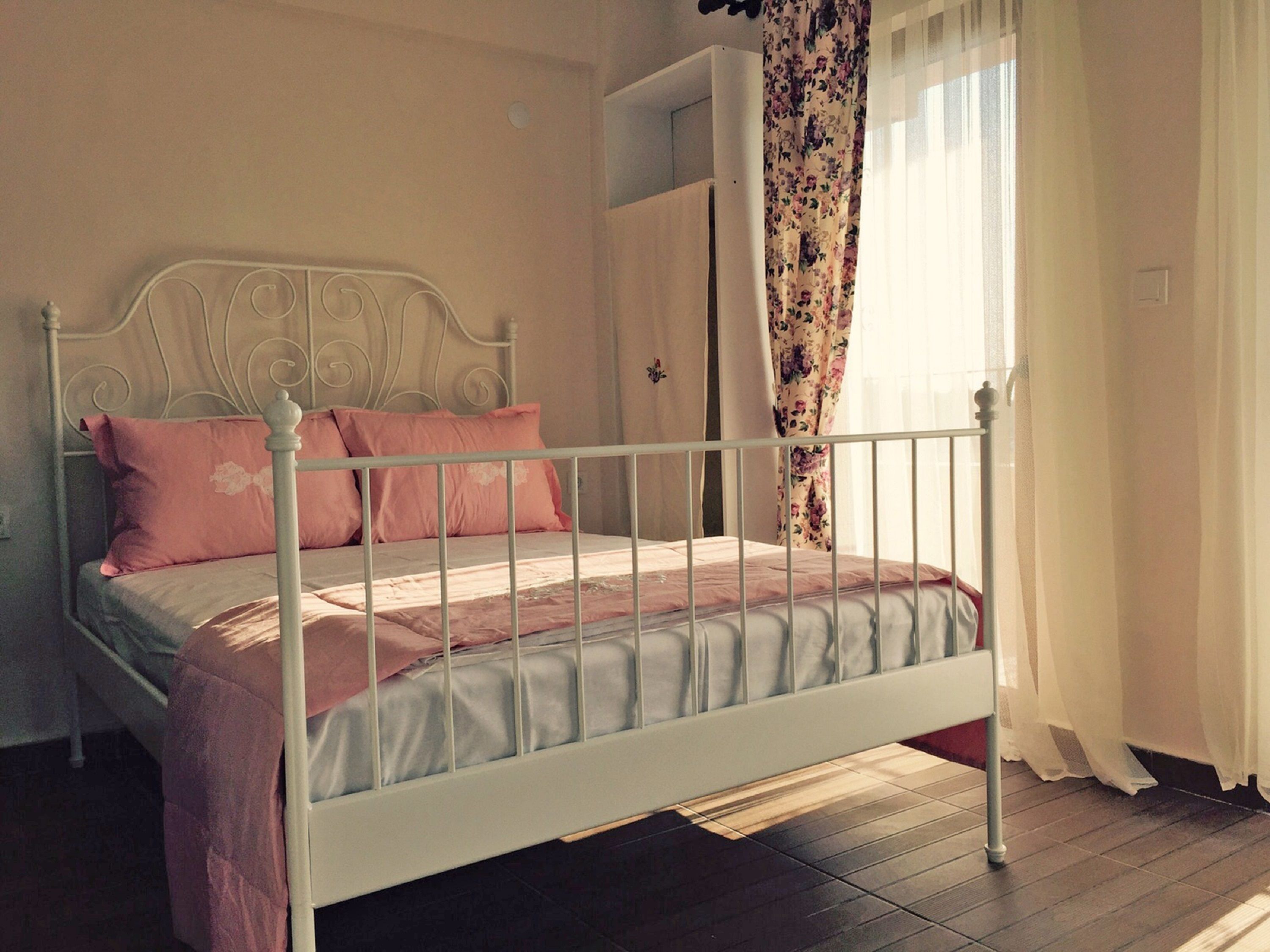 A bedroom with a white metal bed frame, pink and grey bedding, a wooden floor, and a door with floral curtains.