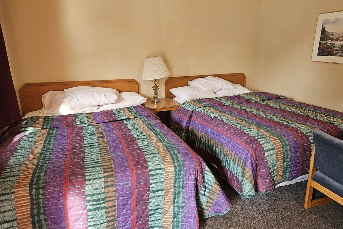 A hotel room with two single beds, each with a colorful quilt, a bedside lamp, and a small table.