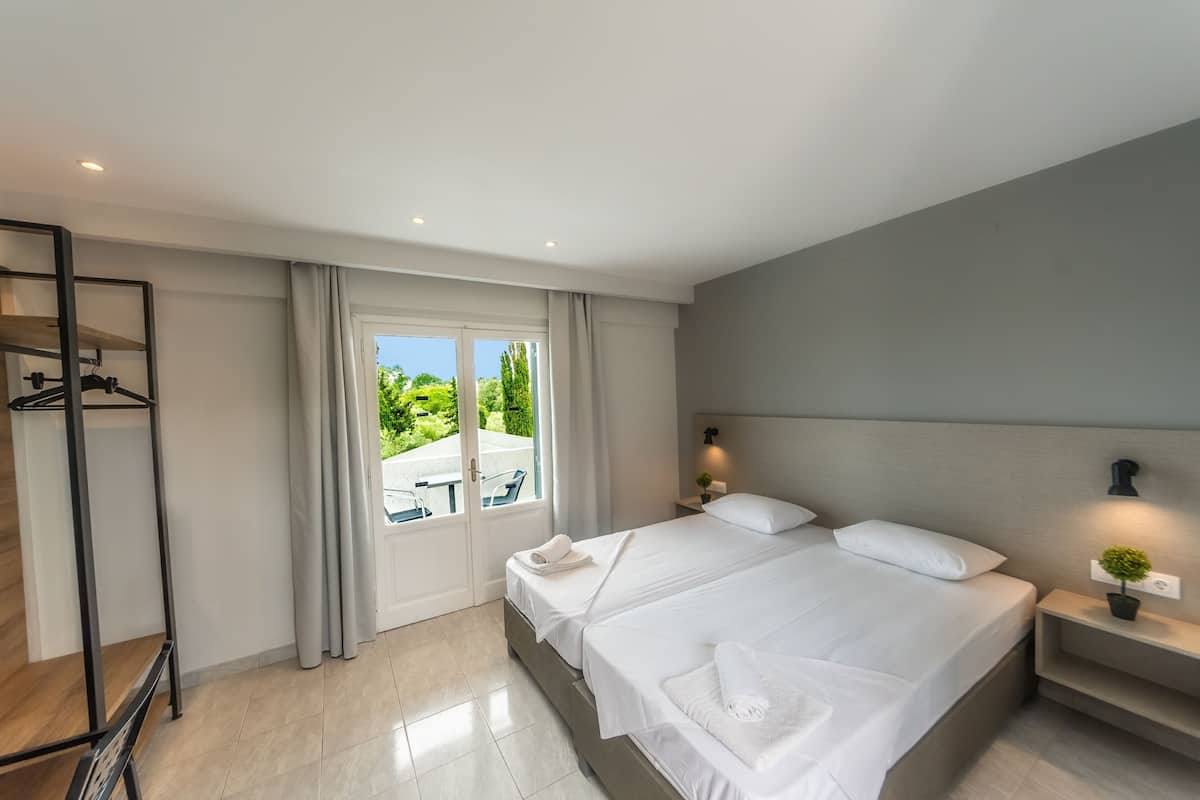 A modern hotel room with a large bed, a bedside table with a lamp, a wardrobe, and a view of the outdoors.