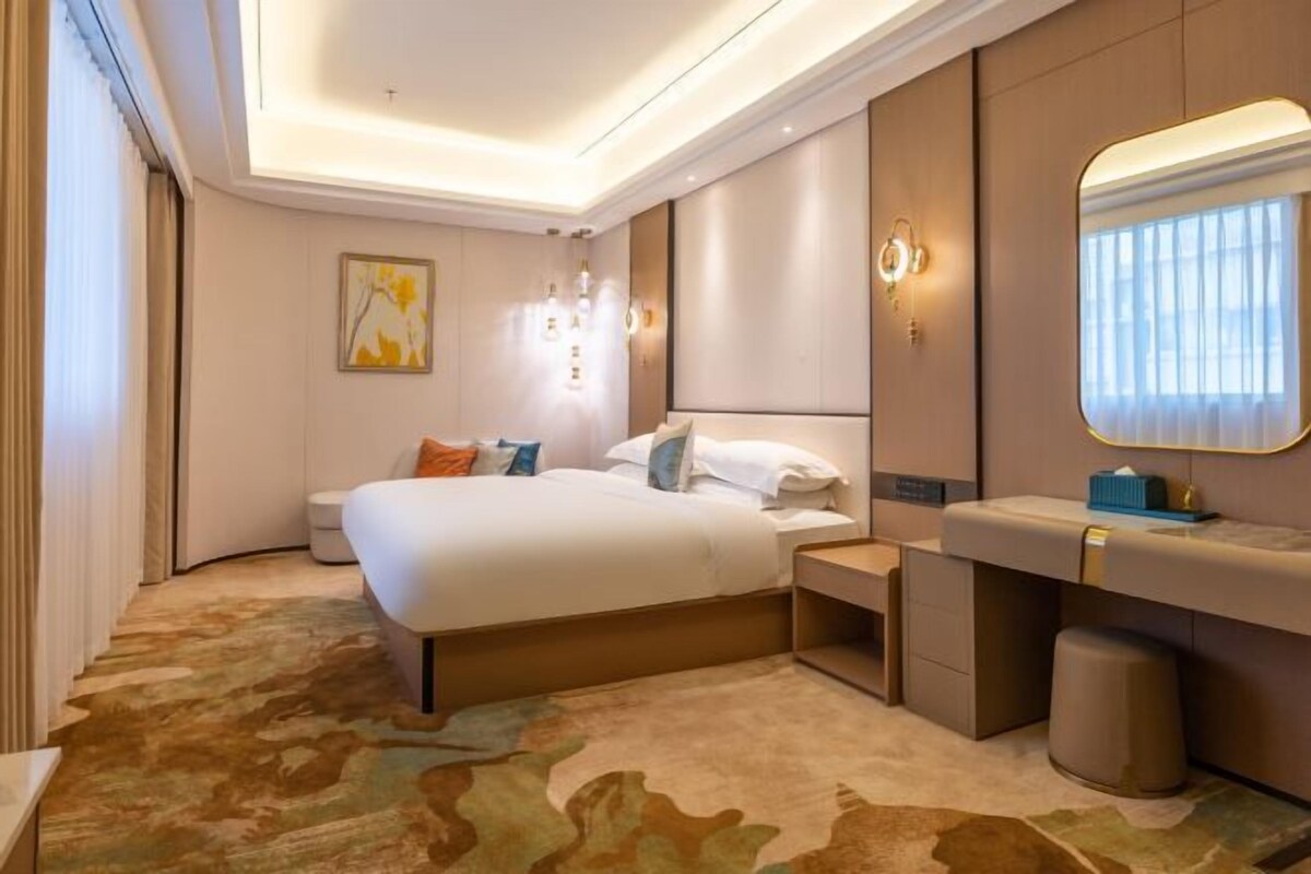 A modern hotel room with a large bed, a bedside table, a mirror, and a carpeted floor.