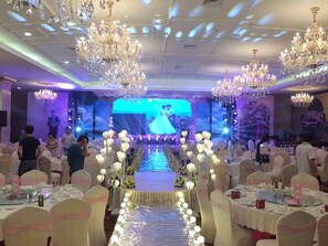 Un salón de banquetes con filas de mesas, candelabros y un escenario con una pantalla grande.
