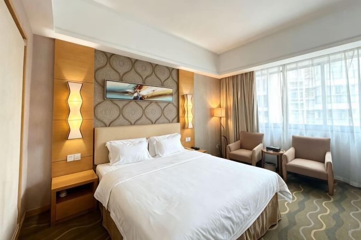 Habitación de hotel con una cama grande, dos sillones, una mesita y un cuadro en la pared, con vistas a la ciudad desde la ventana.