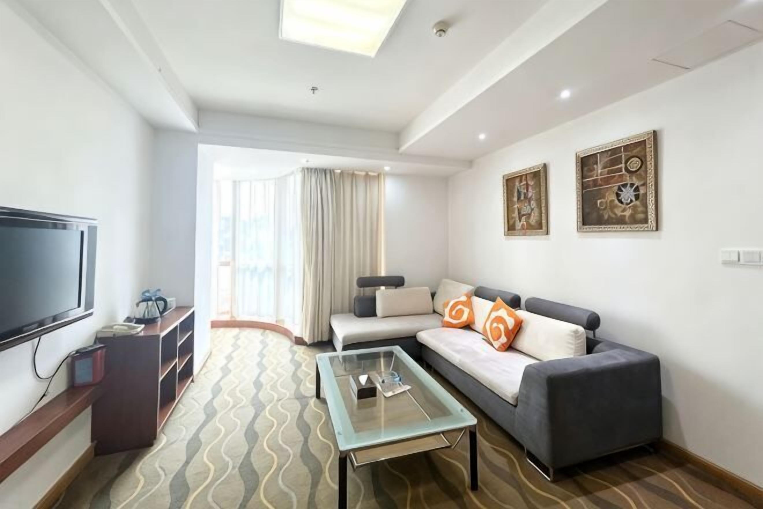 A modern hotel room with a flat-screen TV, a sofa with orange pillows, a glass coffee table, and artwork on the walls.