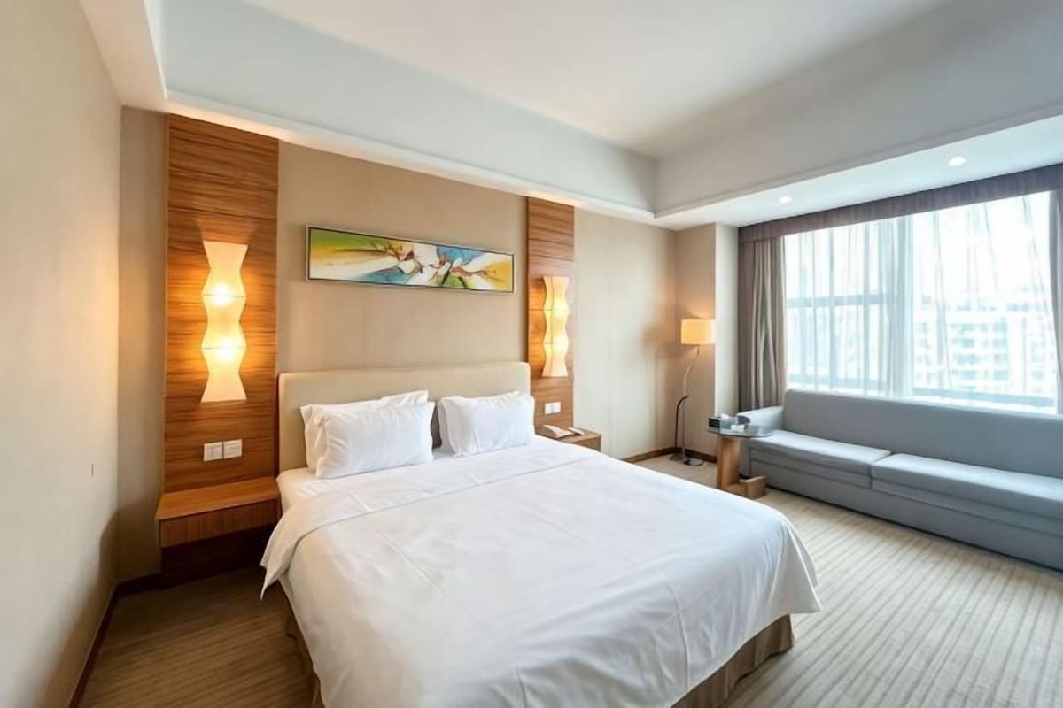 Habitación de hotel con una cama grande, una mesita de noche, una lámpara, una ventana con vistas y un cuadro en la pared.