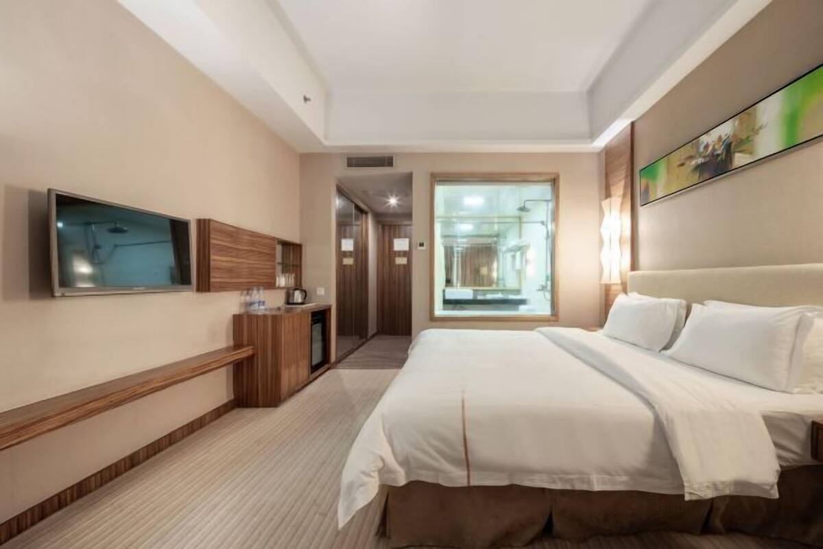 Hotellihuone, jossa on suuri sänky, seinälle asennettu taulutelevisio, puinen työpöytä ja ikkuna, josta on näkymä käytävälle.