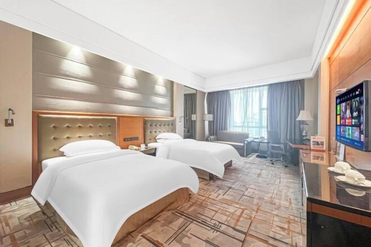 A hotel room with two beds, a desk, a TV, and a large window.