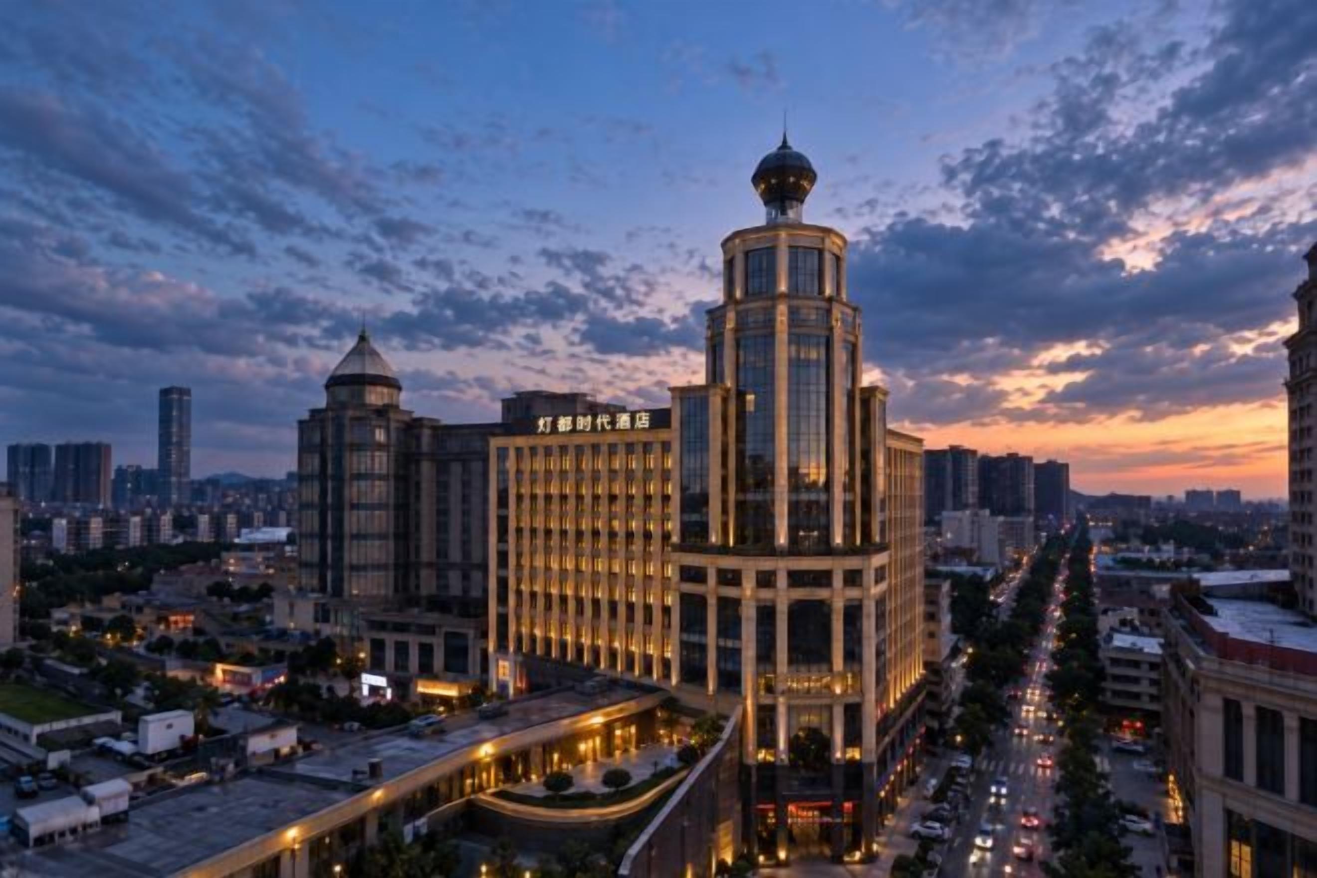 A high-rise building with a prominent central tower and a smaller adjacent structure, surrounded by a cityscape at dusk.