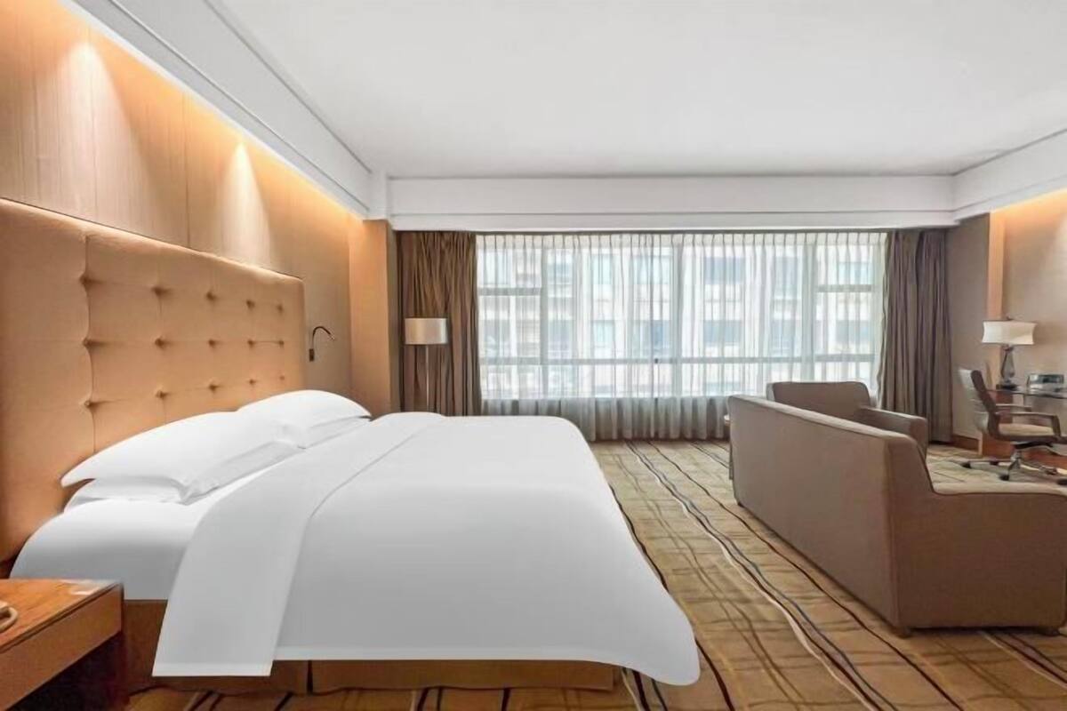 A hotel room with a large bed, a sofa, a desk, and a view of the city through large windows.