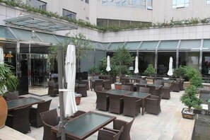 An outdoor patio area with tables and chairs, covered by a glass roof, and surrounded by greenery. - Lighting Era Hotel (Zhongshan)