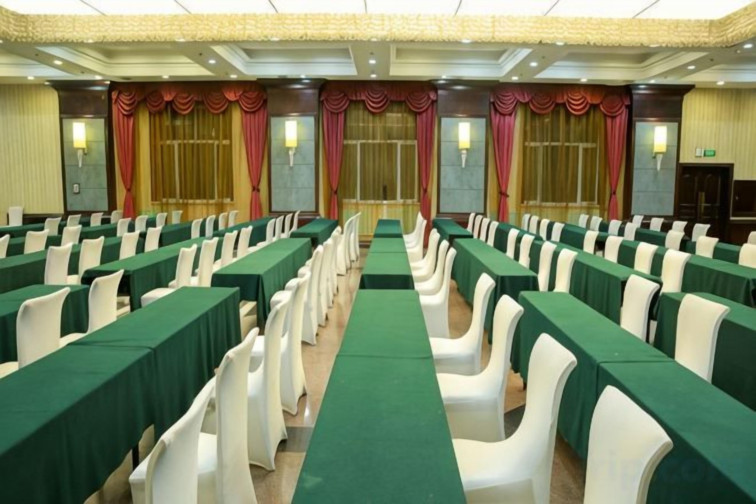 Un salón de conferencias con filas de mesas y sillas, ventanas amplias con cortinas e iluminación empotrada en la pared.