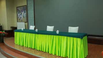 Une table de conférence recouverte d’une nappe verte, avec des chaises blanches et un écran de projection en arrière-plan.