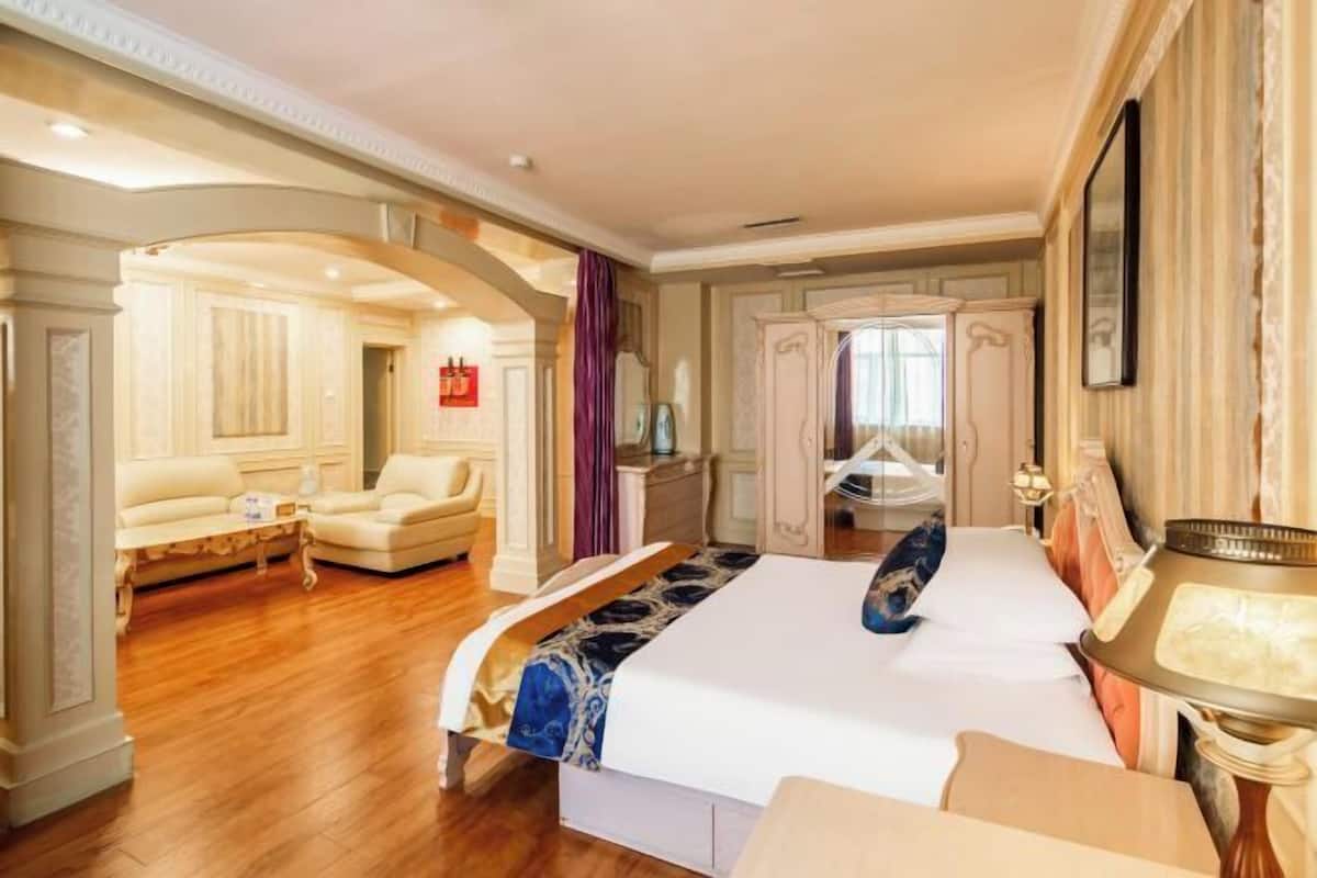 Una habitación de hotel espaciosa con una cama grande, un área de estar con sofá y sillones, y una zona de dormitorio separada con tocador y espejo.