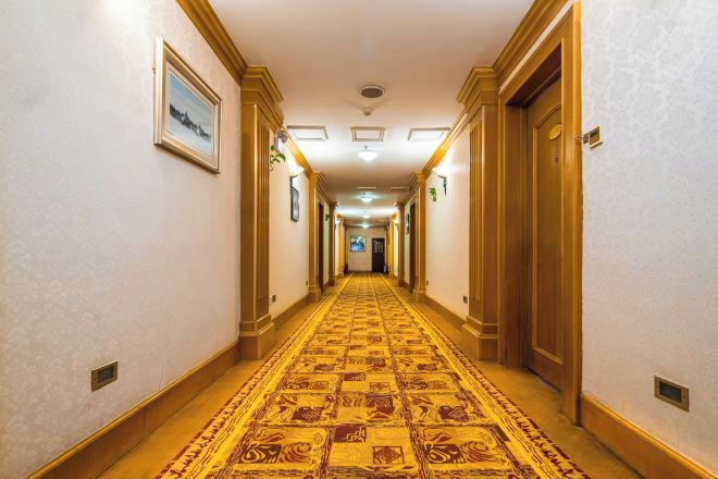 A hotel corridor with patterned carpet, wooden doors, and framed pictures on the walls.