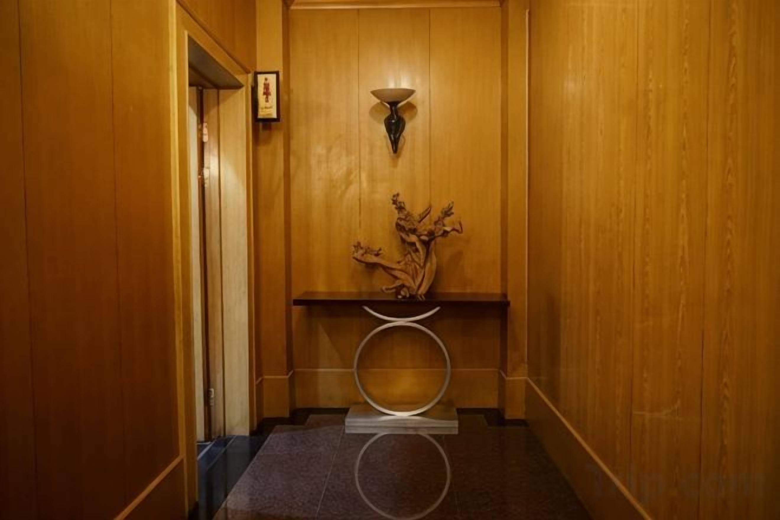 A wooden interior hallway with a door, a wall-mounted light fixture, and a small table with a vase.