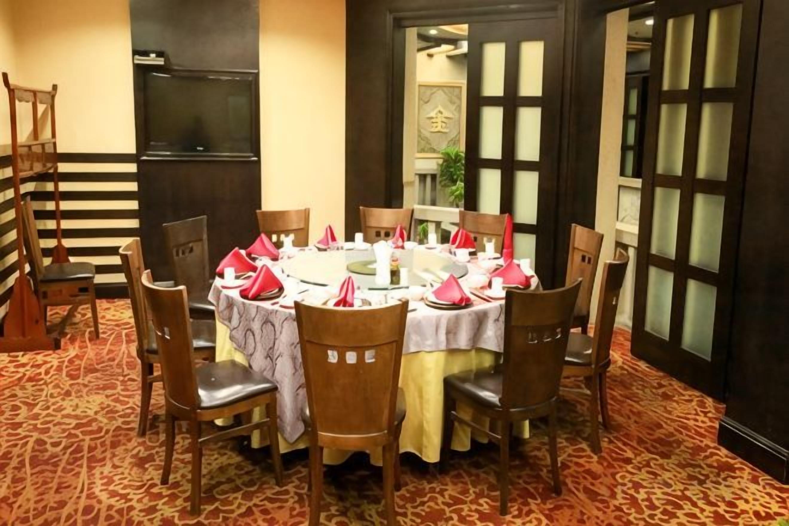 A dining area with a round table set for six, surrounded by wooden chairs, with a carpeted floor and a traditional door in the background.