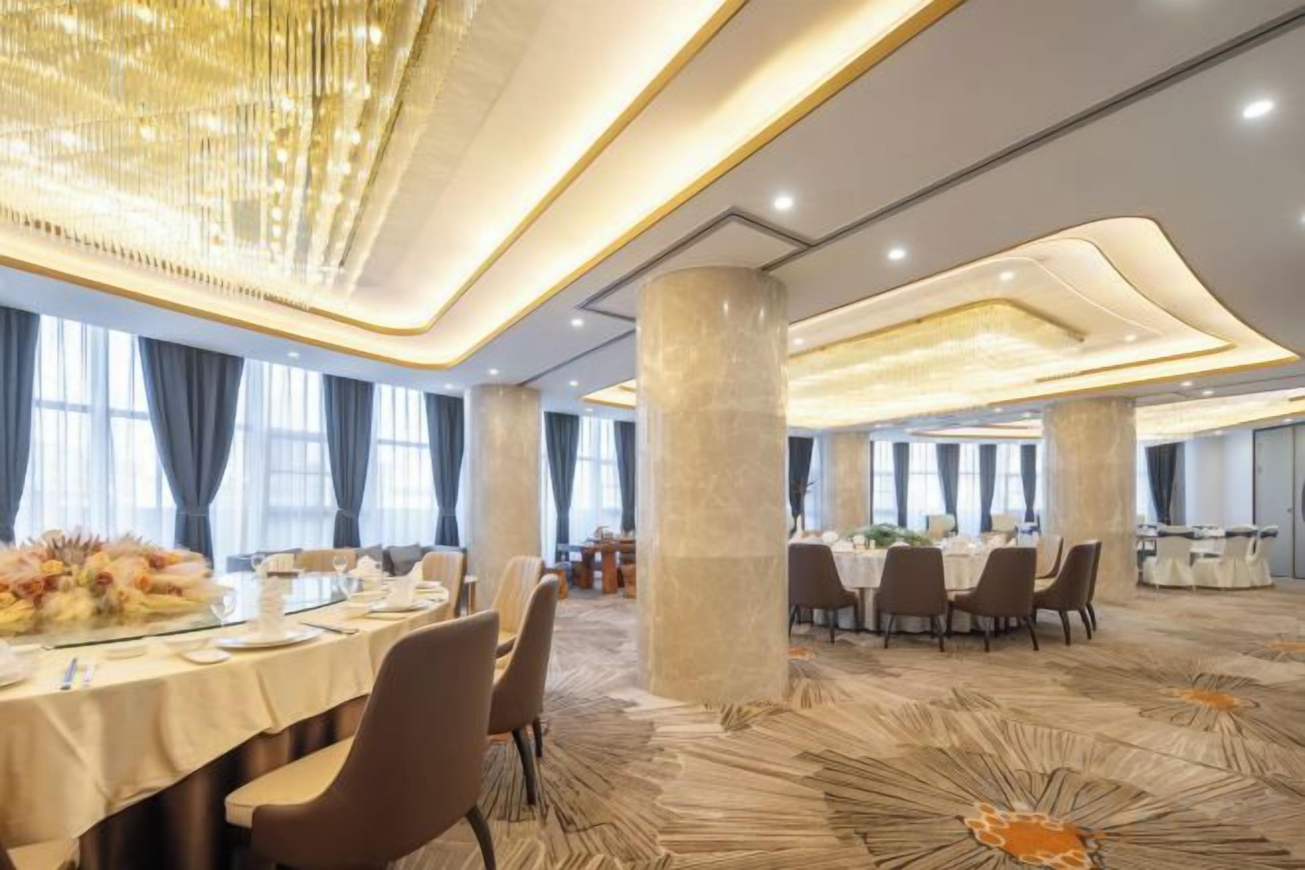 A spacious banquet hall with multiple round tables, large windows, and a modern chandelier.