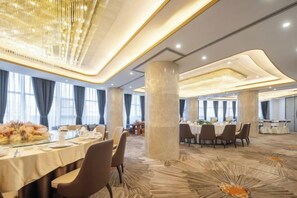 A spacious banquet hall with multiple round tables, large windows, and a modern chandelier.