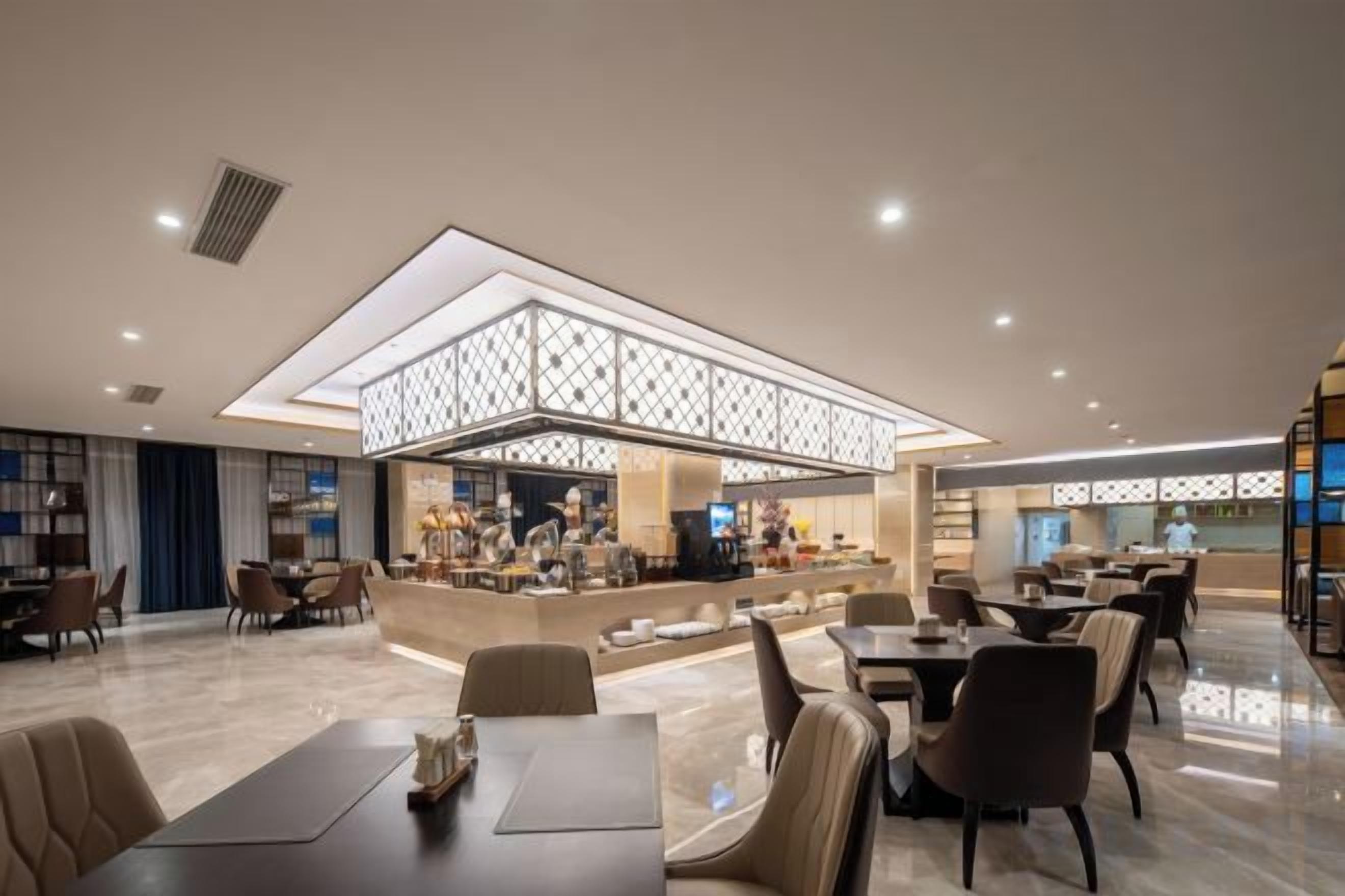 Un hall d’hôtel moderne doté d’un bar central, de différents espaces aménagés pour s’asseoir et d’une structure décorative au plafond.
