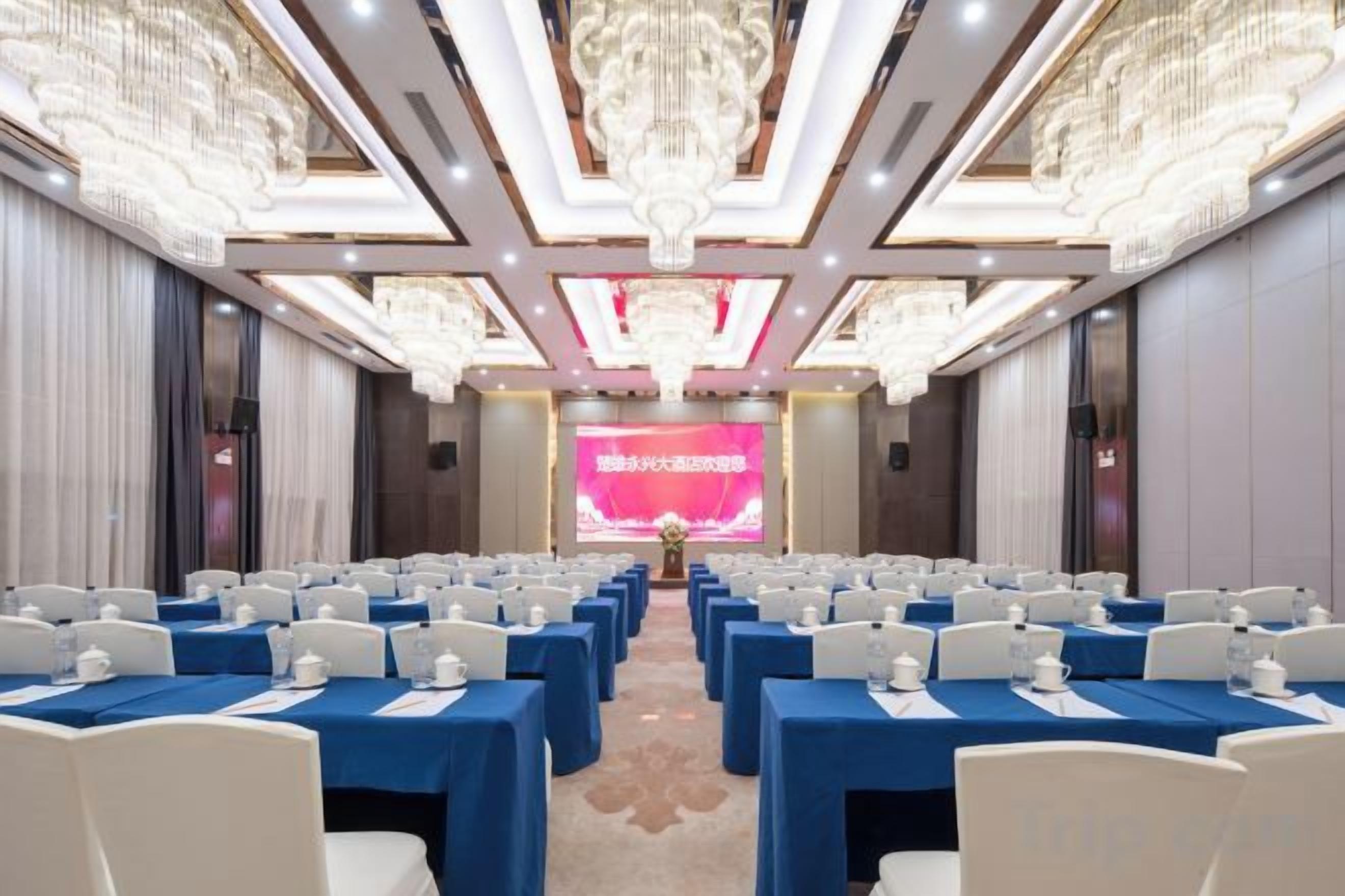 A conference room with rows of tables and chairs, a large screen at the front, and ornate chandeliers.