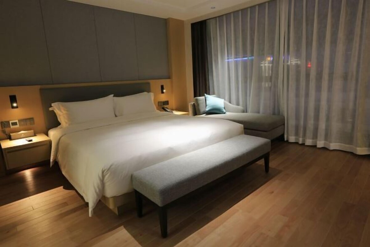 Une chambre d’hôtel moderne avec un grand lit, une banquette, une table de chevet et une fenêtre avec des rideaux.