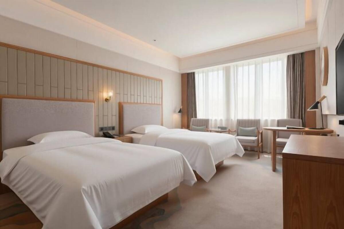A hotel room with two beds, a desk, a chair, and a large window with curtains.