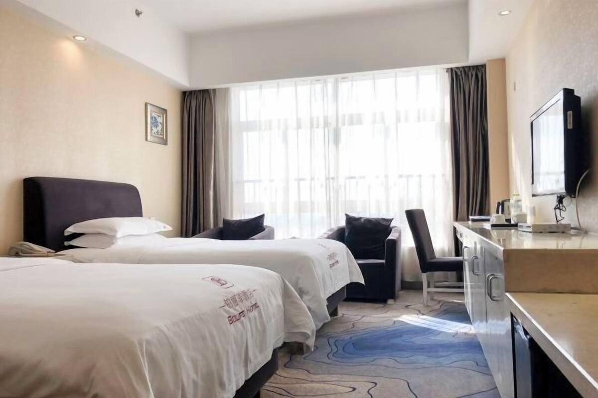 A hotel room with two beds, a desk, a chair, a TV, and a large window with curtains.