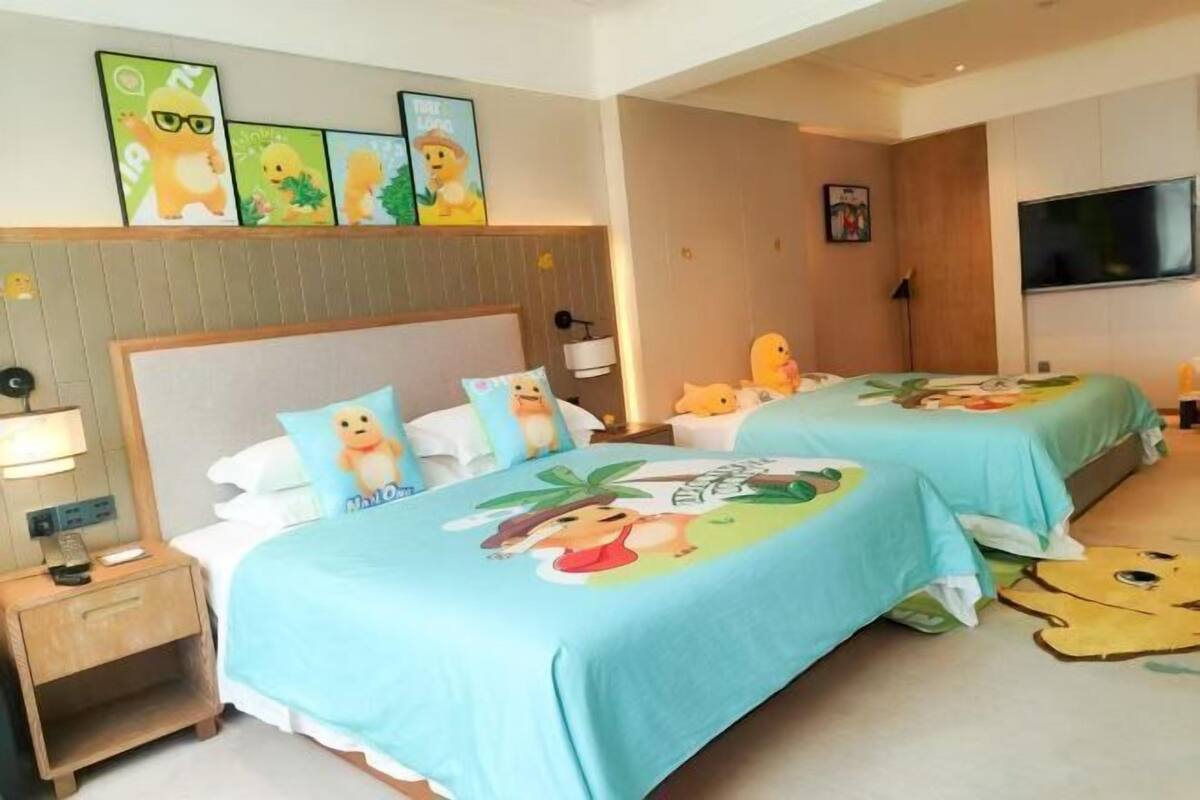 A hotel room with two beds, each with a blue bedspread featuring cartoon characters, a wooden headboard, a bedside table with a lamp, a TV mounted on the wall, and a framed picture above the beds.