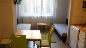 A small, compact room with two single beds, a small table, a chair, a radiator, and a kitchenette. - Alisa Apartments (Kostomlaty nad Labem)