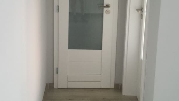 A narrow hallway with a white door at the end, a small step leading up to it, and a light switch on the wall to the right. - Alisa Apartments (Kostomlaty nad Labem)