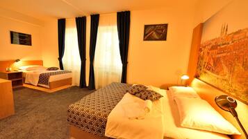 A hotel room with a large bed, a smaller bed, a desk, and a chair. There are curtains, a painting on the wall, and a lamp on the desk.