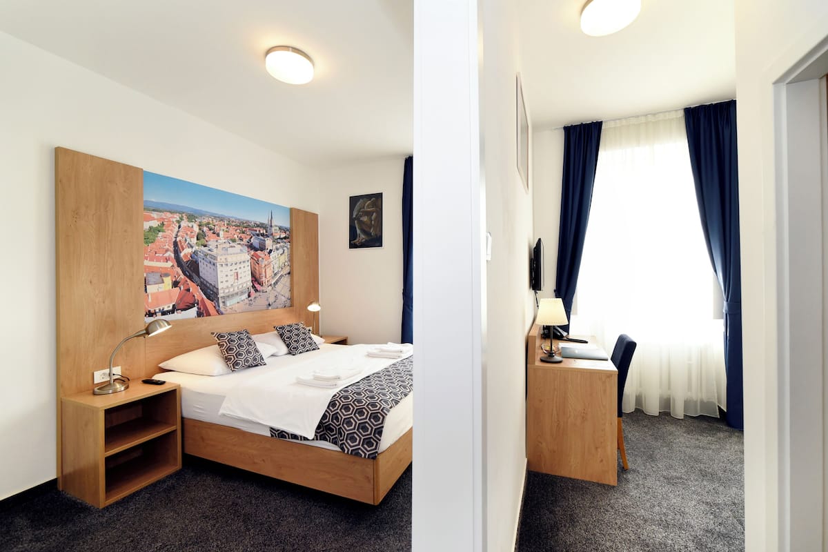 Una habitación de hotel con cama, mesita de noche, lámpara y un cuadro de un paisaje urbano.
