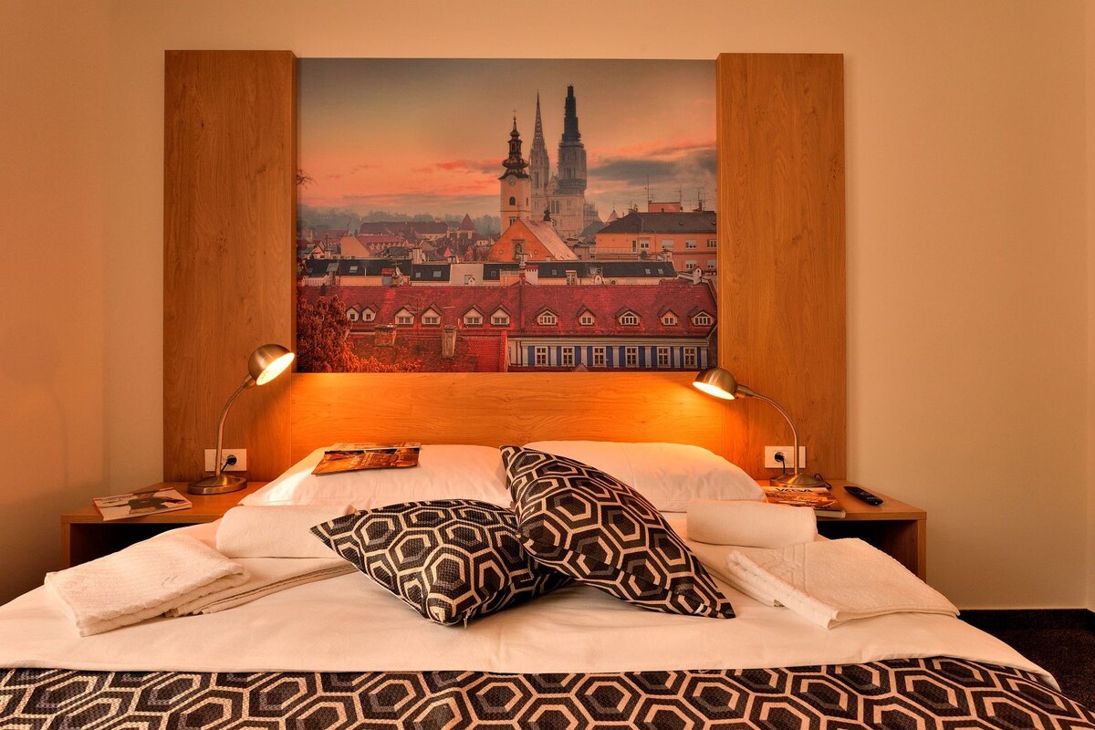 A hotel room with a large bed, two bedside lamps, a headboard with a cityscape mural, and a neatly made bed with patterned pillows.