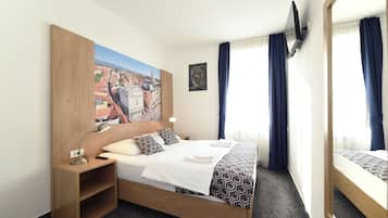 Habitación de hotel con cama, mesita de noche, un cuadro, televisor y ventana con cortinas.