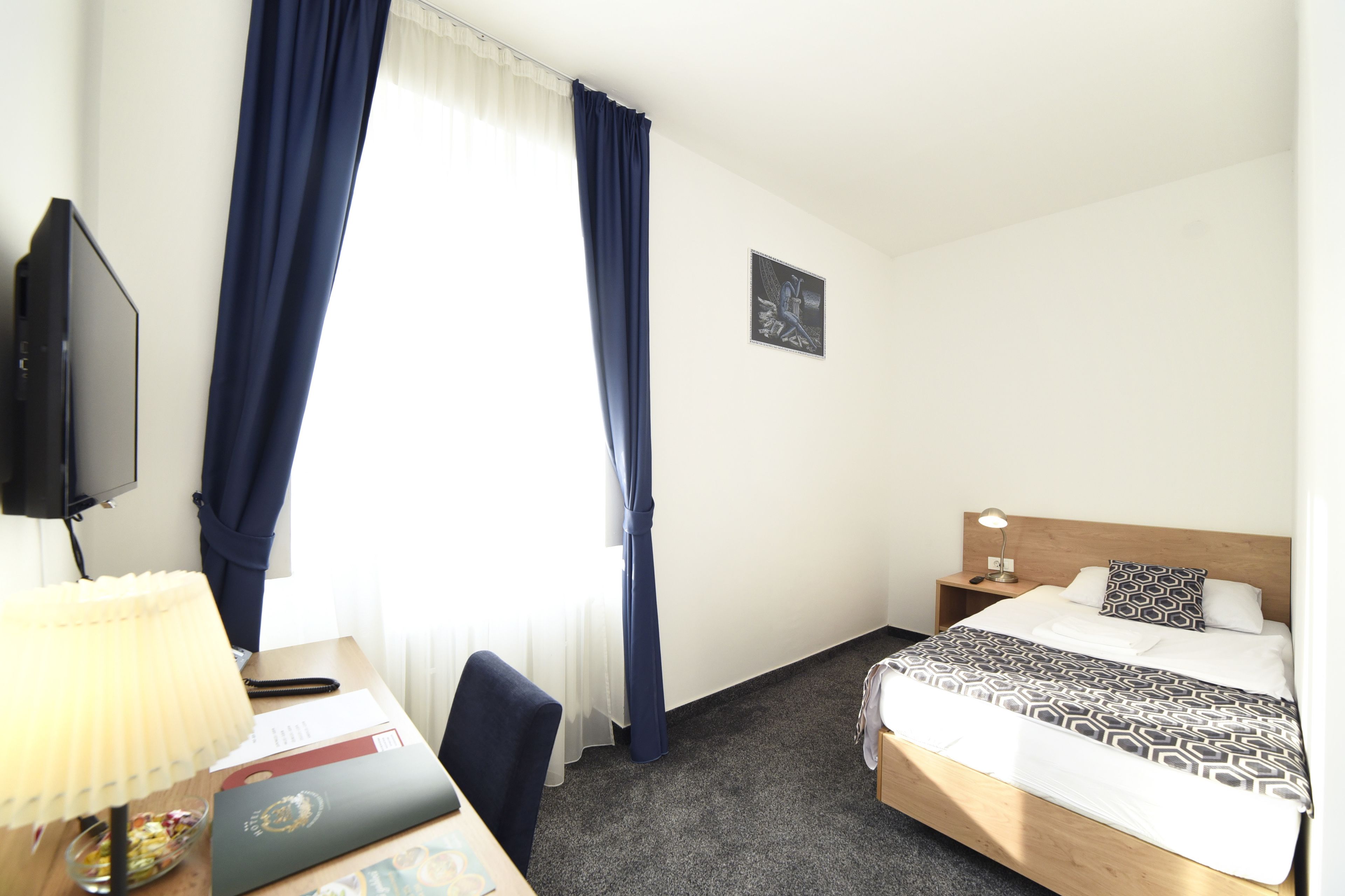 A hotel room with a bed, a desk with a lamp, a chair, a television, and a window with curtains.