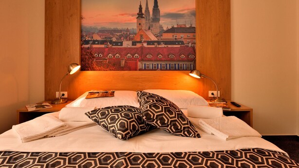 A hotel room with a large bed, two bedside lamps, a headboard with a cityscape mural, and a wooden headboard. - Hotel Orient Zagreb (Zagreb)
