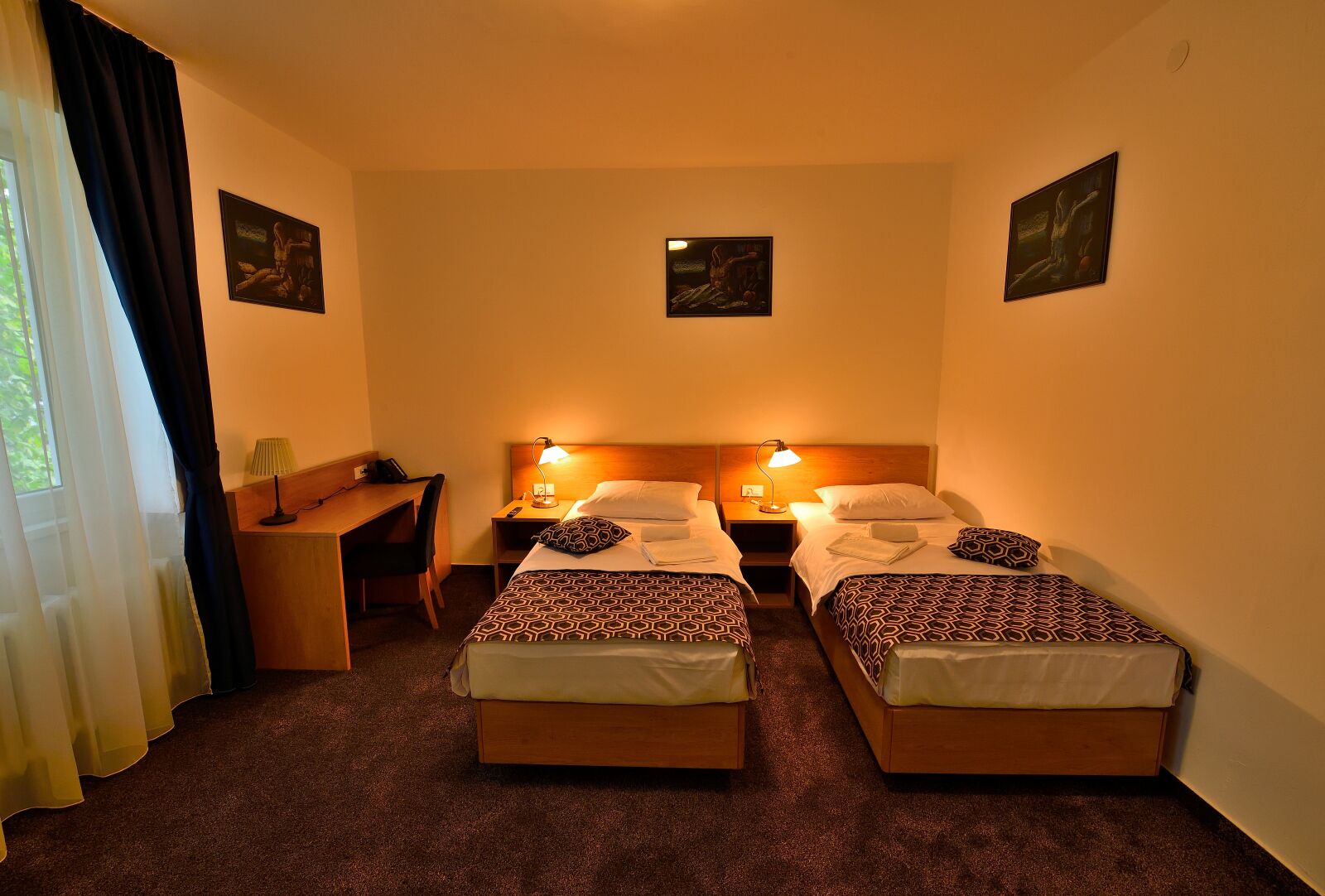 Ein Hotelzimmer mit zwei Einzelbetten, einem Schreibtisch, einem Stuhl und einem kleinen Tisch mit einer Lampe.