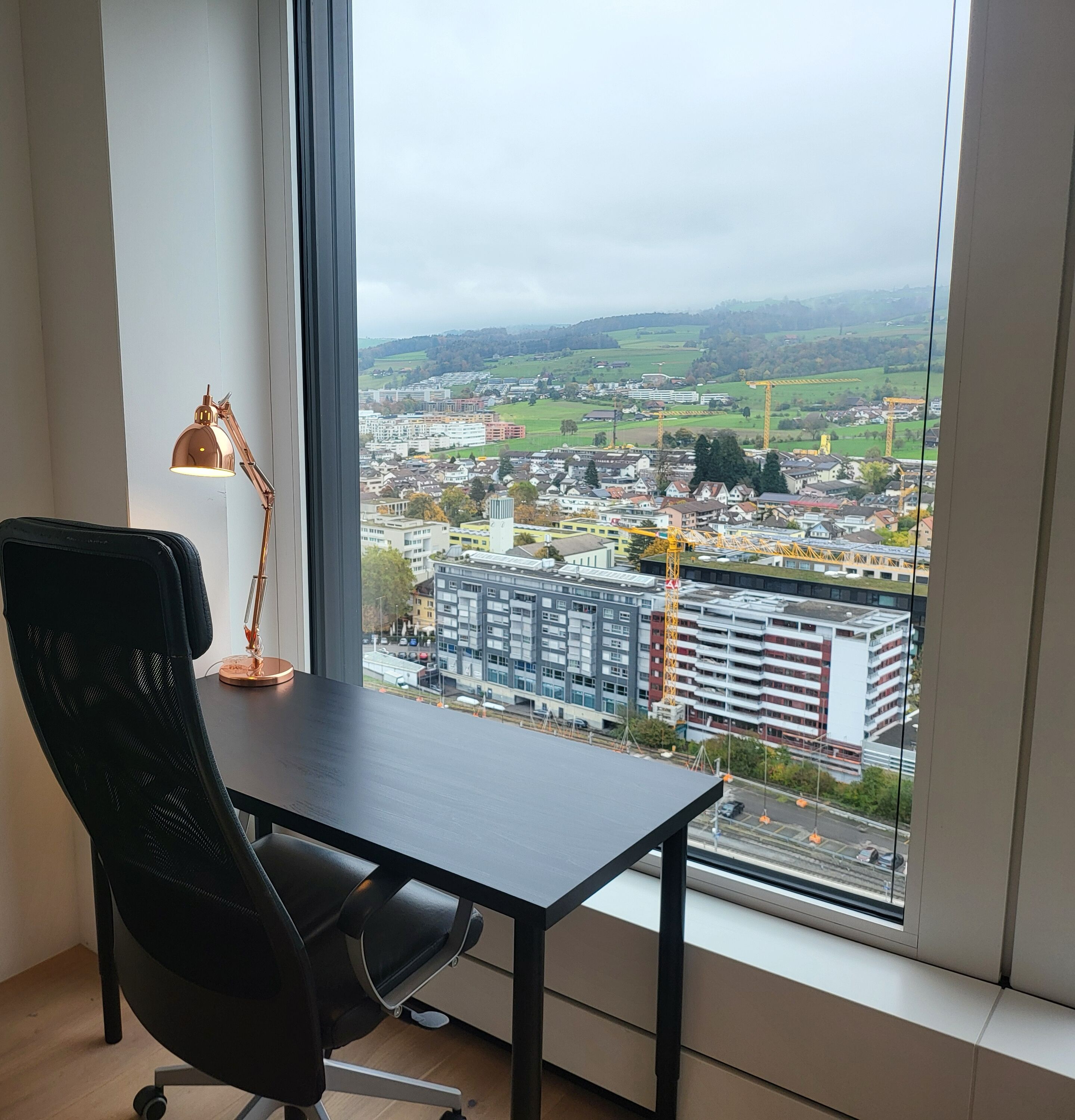 A room with a desk, chair, and a large window offering a city view.