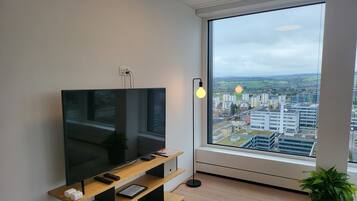 A modern living room with a flat-screen TV on a wooden stand, a floor lamp, and a large window offering a city view.