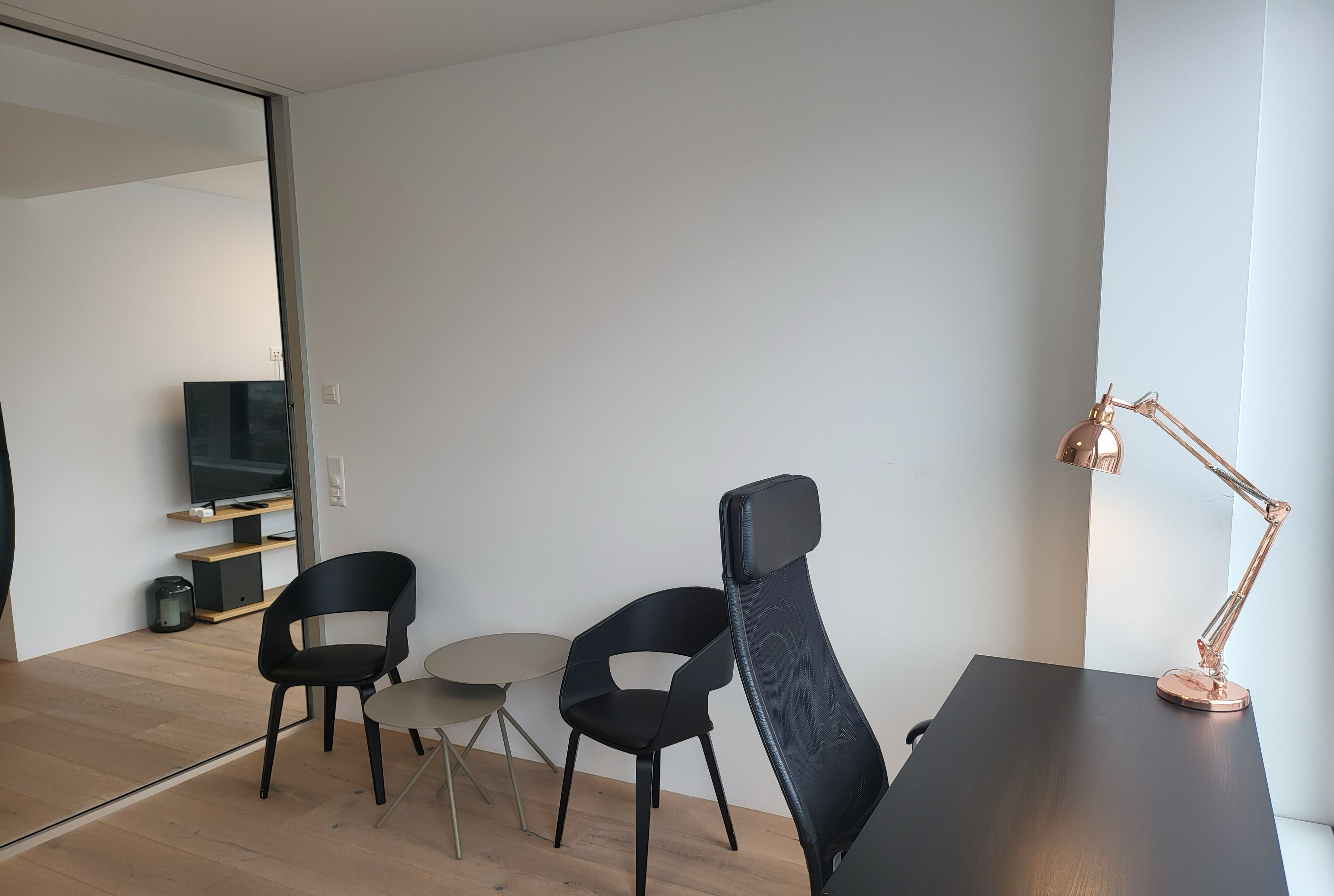 A modern, minimalist room with a dark wooden desk, two black chairs, a small round table, and a single table lamp. A television is mounted on a wooden shelf, and a sliding door leads to another room.