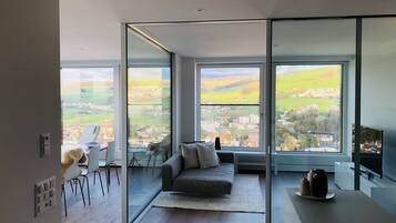 A modern living room with a sofa, a coffee table, and a TV. Large windows offer a view of a cityscape.