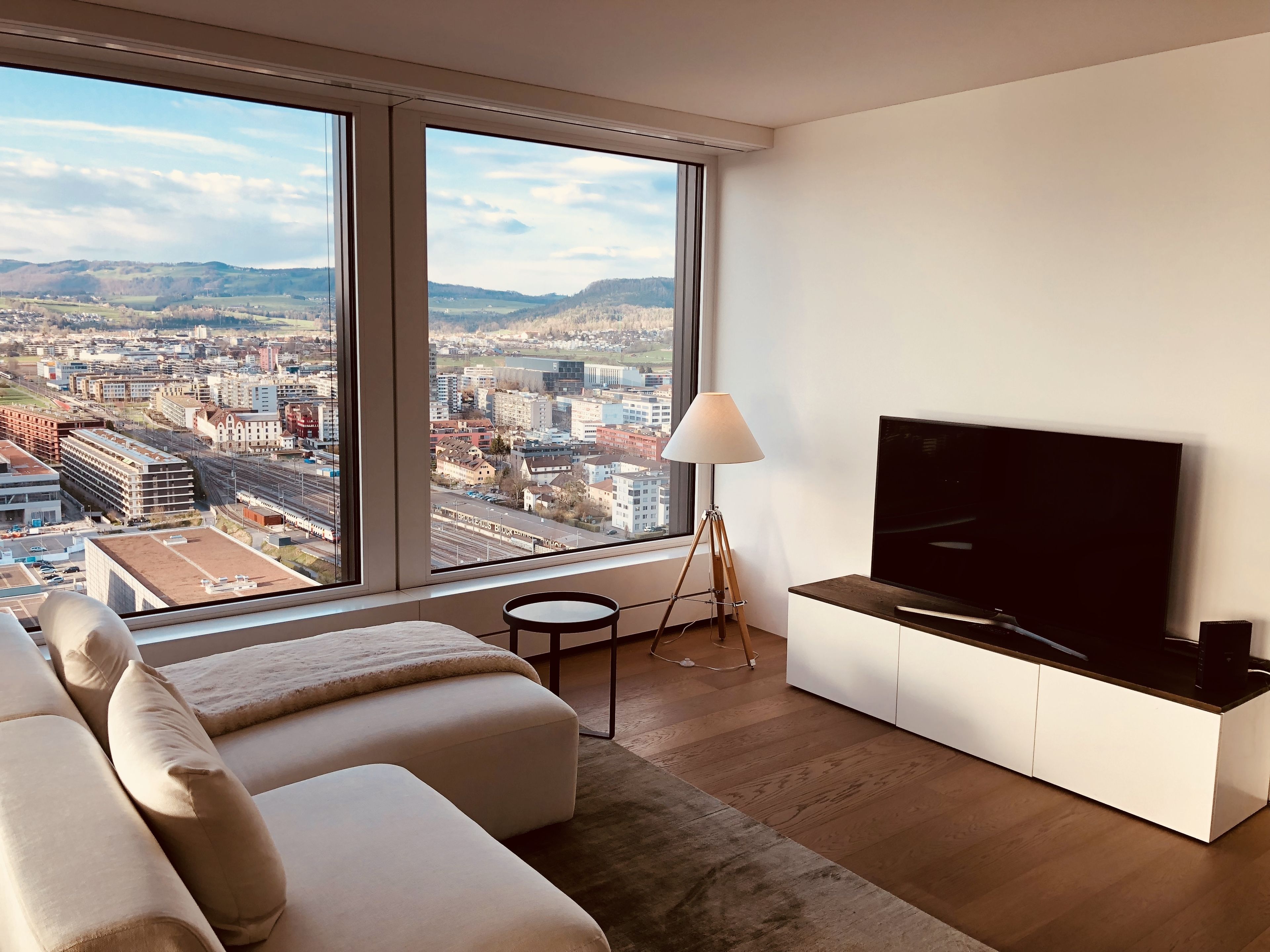 A modern living room with a large window, a flat-screen TV, a sofa, and a lamp.