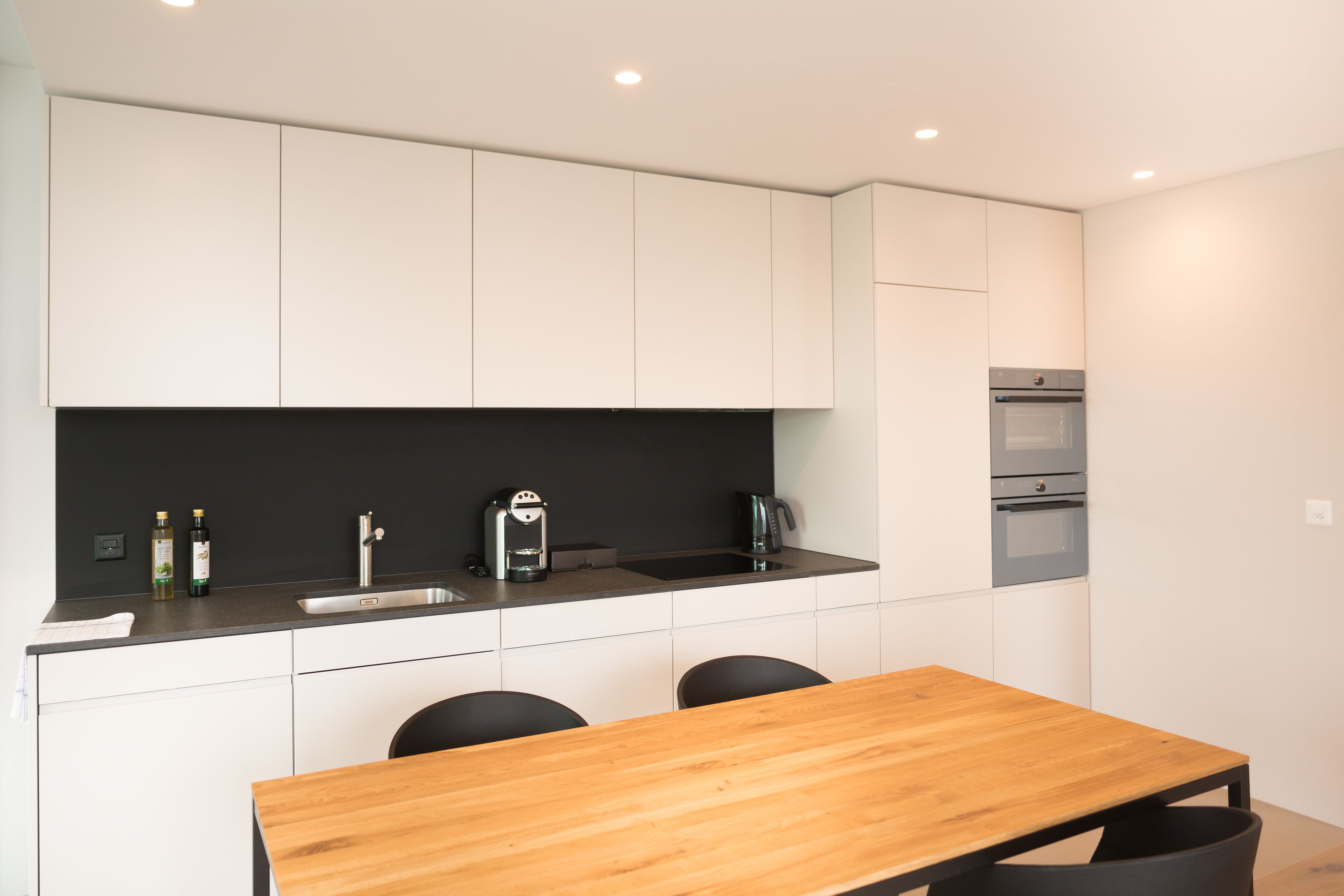 A modern kitchen with white cabinets, a black countertop, a wooden table, and black chairs.