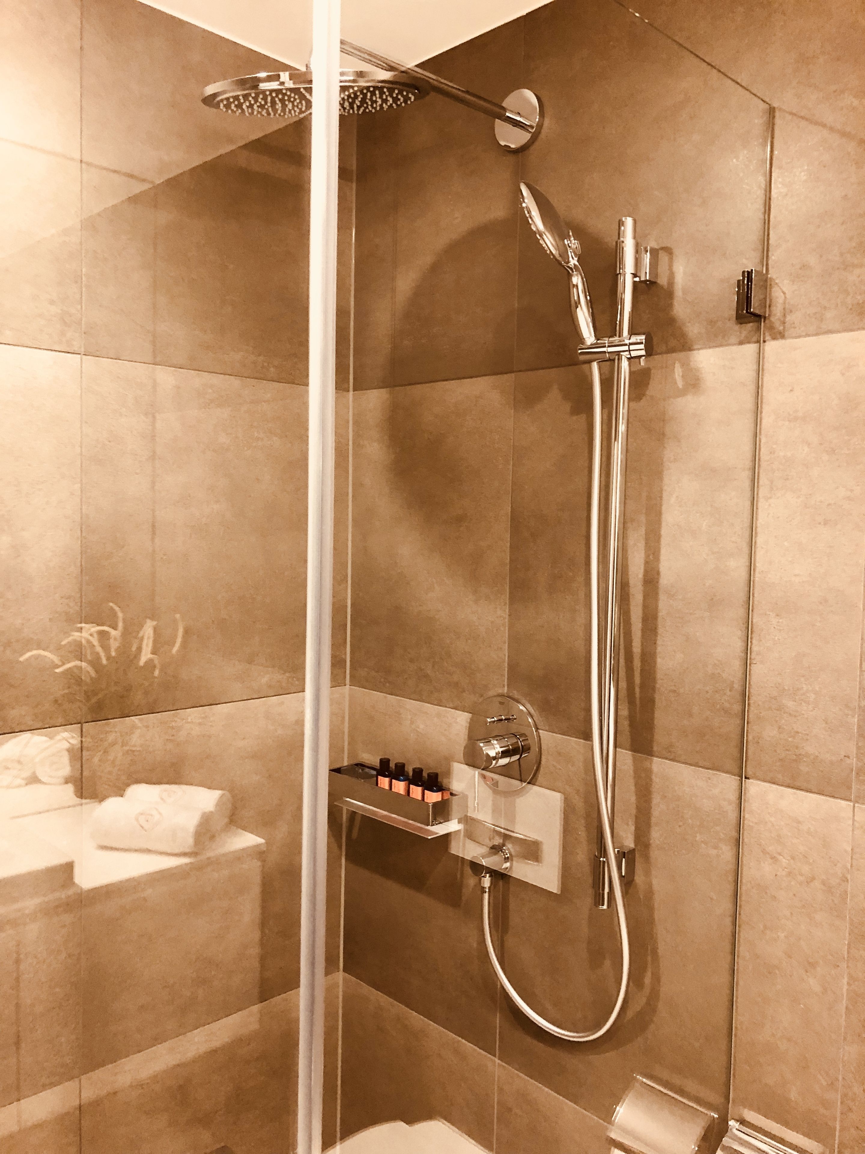 A modern shower with a handheld showerhead and a wall-mounted faucet, a glass-enclosed shower area, and a shelf with toiletries.